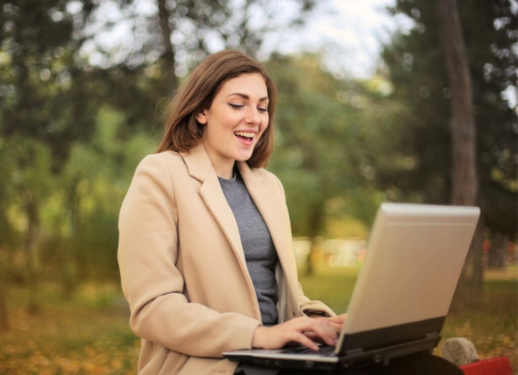 Professional woman in beige coat and gray sweater smiling while working on laptop in natural outdoor setting